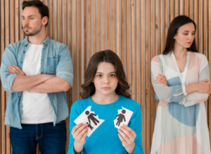 Sad child holding torn images of a family with parents in the background.